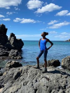 Diana standing on a rock in front of the Pacific ocean in a blue tank top and black shorts