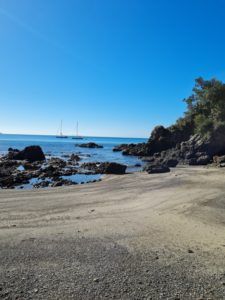 Sandy beach with black rocky outcrop and two sail boats off in the distance. clear blue skies